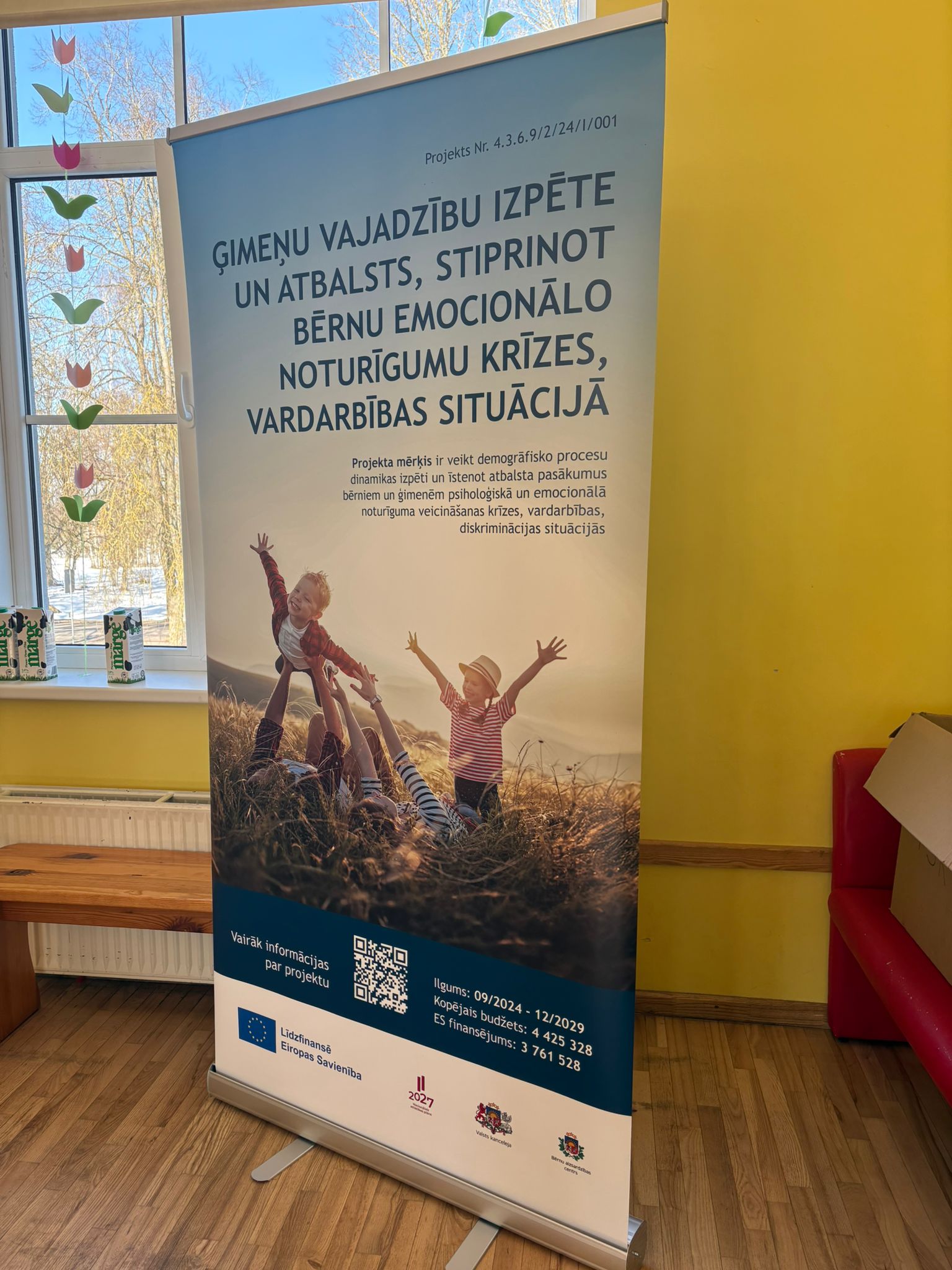 **ALT teksts (mājaslapai):** Roll-up stends Vecsaules pamatskolas telpās ar informāciju par projektu par ģimeņu vajadzību izpēti un atbalstu bērnu emocionālās noturības stiprināšanai krīzes un vardarbības situācijās. **Nedaudz aprakstošāks ALT teksts (ja vajag pieejamībai):** Vecsaules pamatskolas telpā novietots roll-up stends ar projekta informāciju par ģimeņu vajadzību izpēti un atbalstu bērnu emocionālās noturības stiprināšanai krīzes, vardarbības un diskriminācijas situācijās. Ja vēlies, varu arī uzrakstīt **SEO optimizētu attēla aprakstu mājaslapai**, kas palīdz labāk parādīties Google rezultātos.