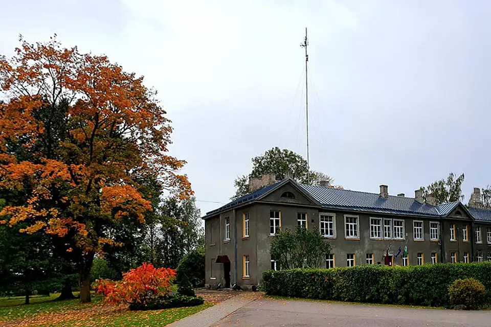 Skolas foto - skolas ēka. Fonā zaļi koki, zāle. Akmens ar skolas uzrakstu